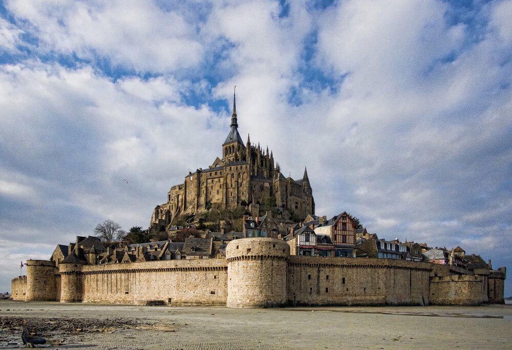 Visiting le Mont Saint Michel| soaring spire on a church on an island; fortified wall surrounds it