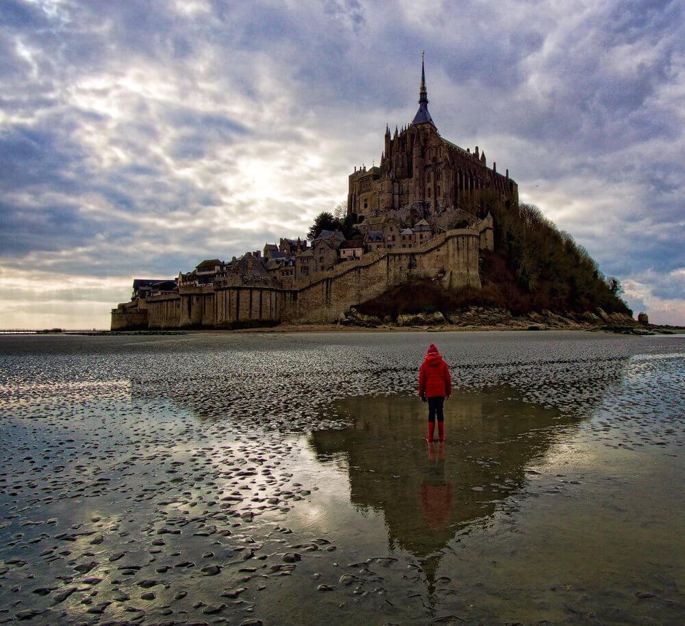 Visit Mont-St-Michel | child in red coat standing in a puddle's reflection of the Abbey