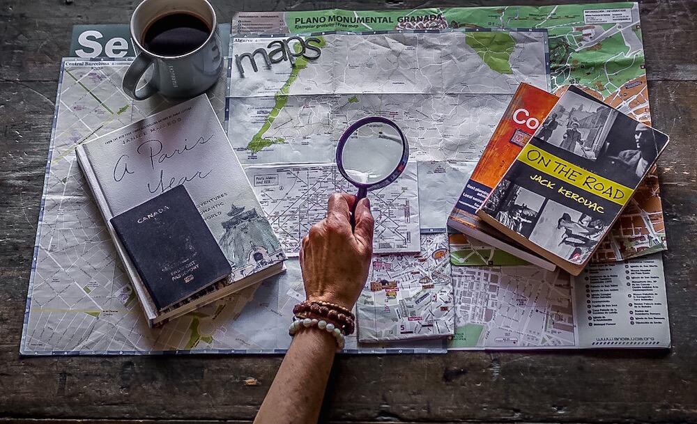 plan my journey: maps, travel books stacked. hand holding magnifying glass