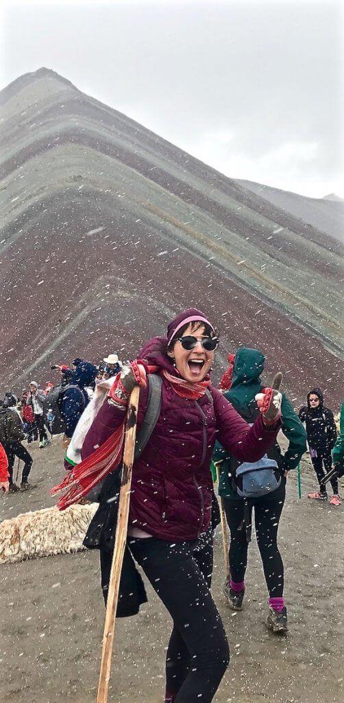 lady in maroon hat and jacket with walking stick, in a blizzard with the stripes of rainbow mountain behind muted colours due to the snow