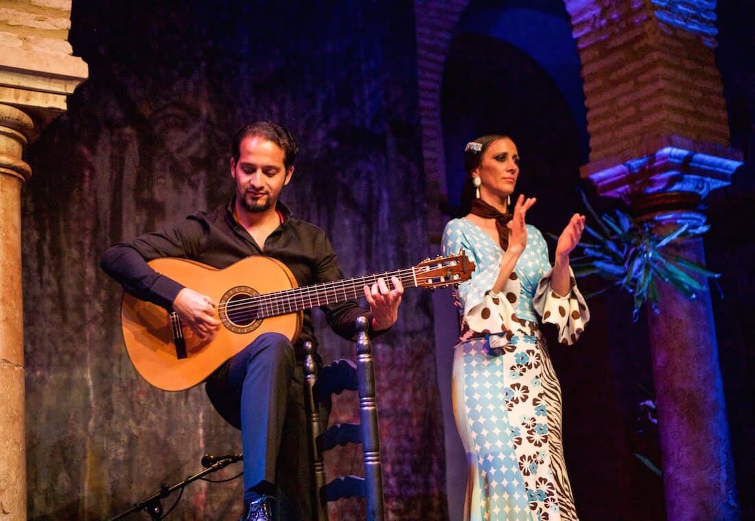 Seville has flamenco dancing. A man playing the guitar and a lady clapping.