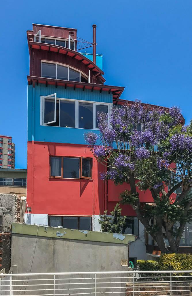 Things to do in Valparaiso: this tall house that is red and blue with a rounded staircase outside at the top, window open, flowering tree in front
