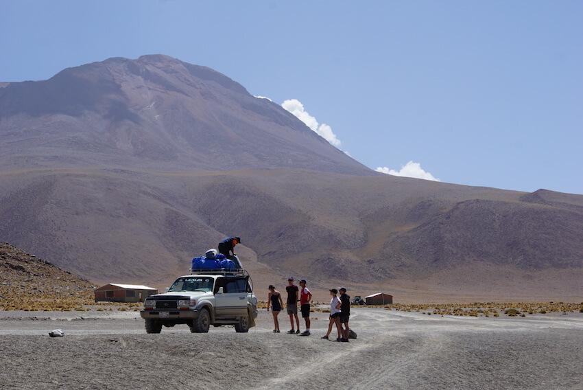 The white jeep in front of the Andes; 5 people standing around; leader getting gas off the roof