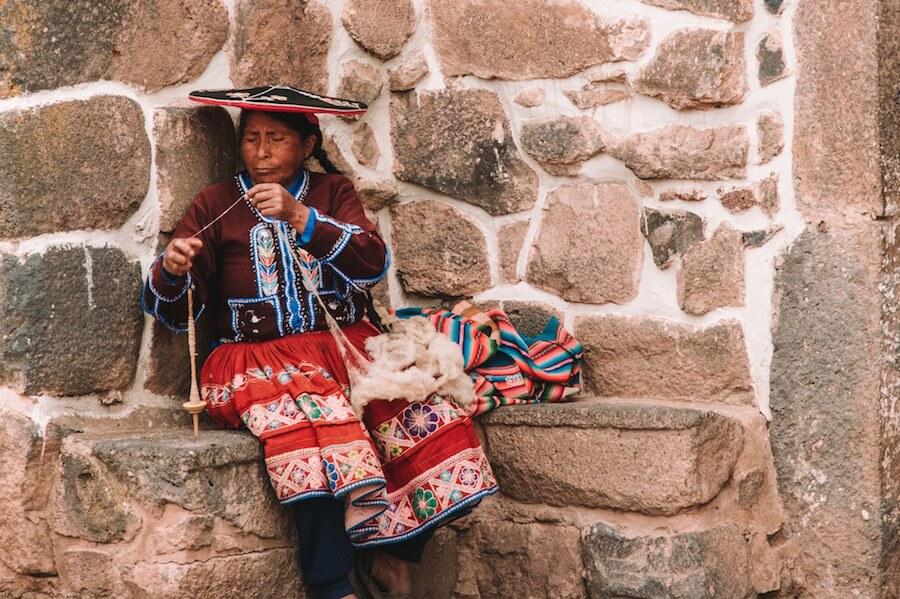 Visit Peru and see old ways of life in action. Lady in flat hat weaving alpaca wool from a spindle. Stone wall behind her.