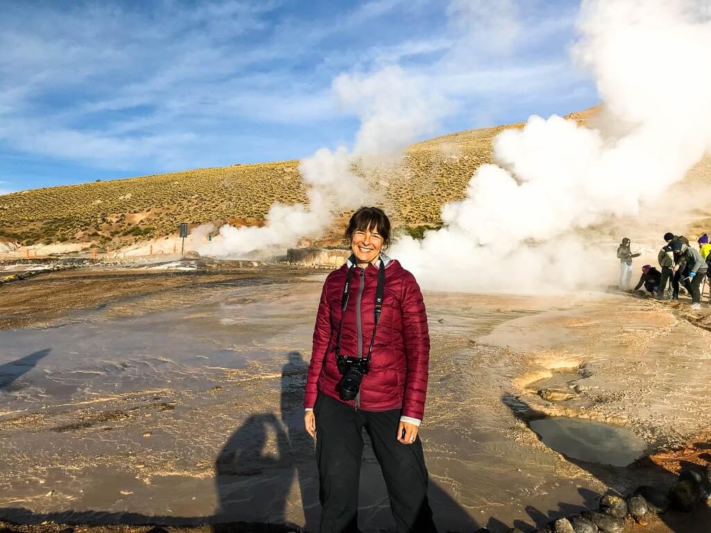 Lady standing in burgundy jacket, geysers steaming behind