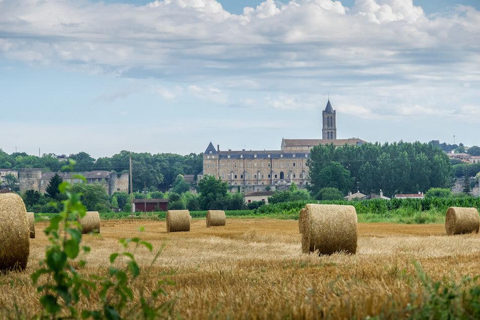 hay bales rolled in the foreground, green trees and a rectangular brick building with a tower , its the abbey at La Reole