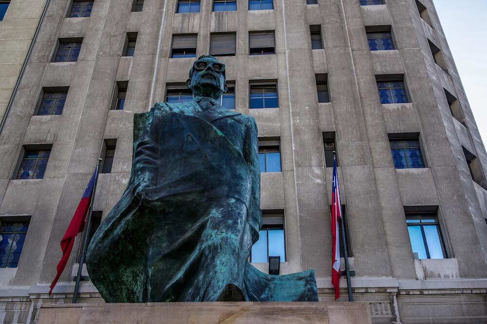 Things to do in Santiago Chile| see the green statue of a Salvador Allende wearing glasses, Chilean flags on the building behind flank the statue