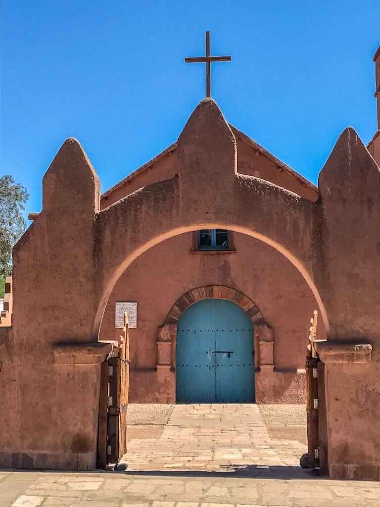 Red adobe chirch with arch in front, blue door and simple cross against a blue sky