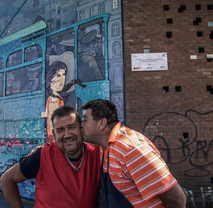 One man kissing the other on the cheek; horsing around for the camera, street art in the background