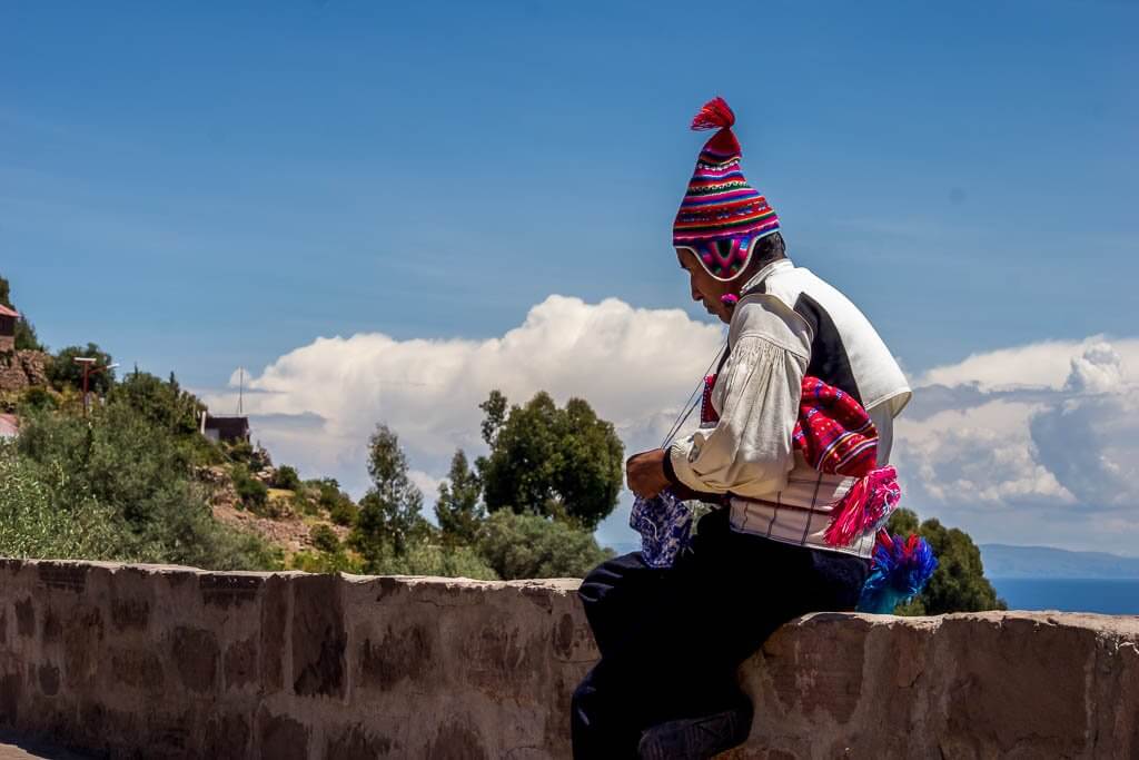 South America Travel route: man knitting sitting on stone wall, Lake titicaca in background. He is wearing traditional knitted hat, mostly red. with tassel at top