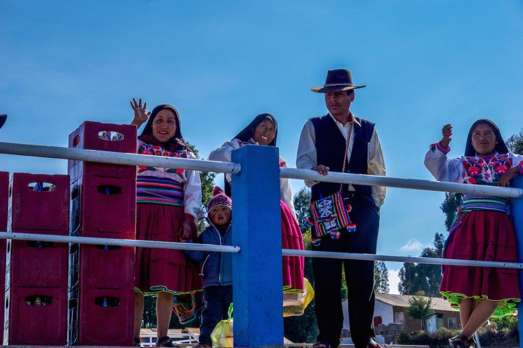 South America Travel route: 3 woman and a man bid farewell to leaving tourists. They wear traditional dress, Women in red skirts, embroidered blouses and black head scarves