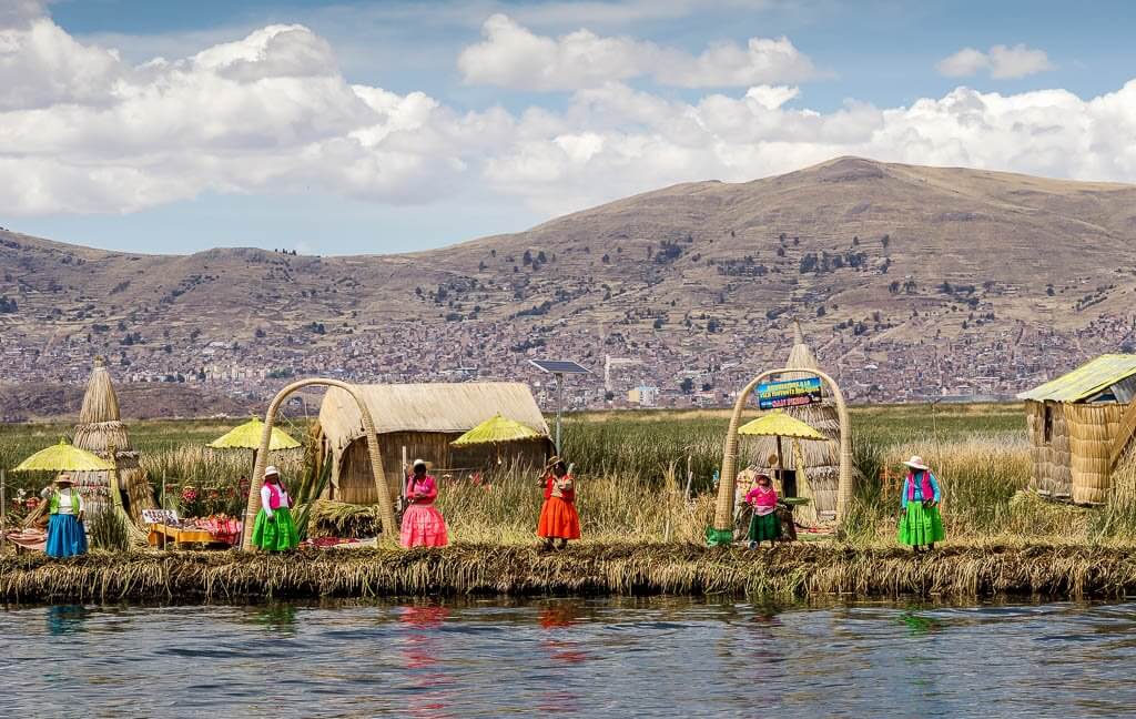 Uros: ladies line up along shore wearing brightly coloured skirts with reed huts behind