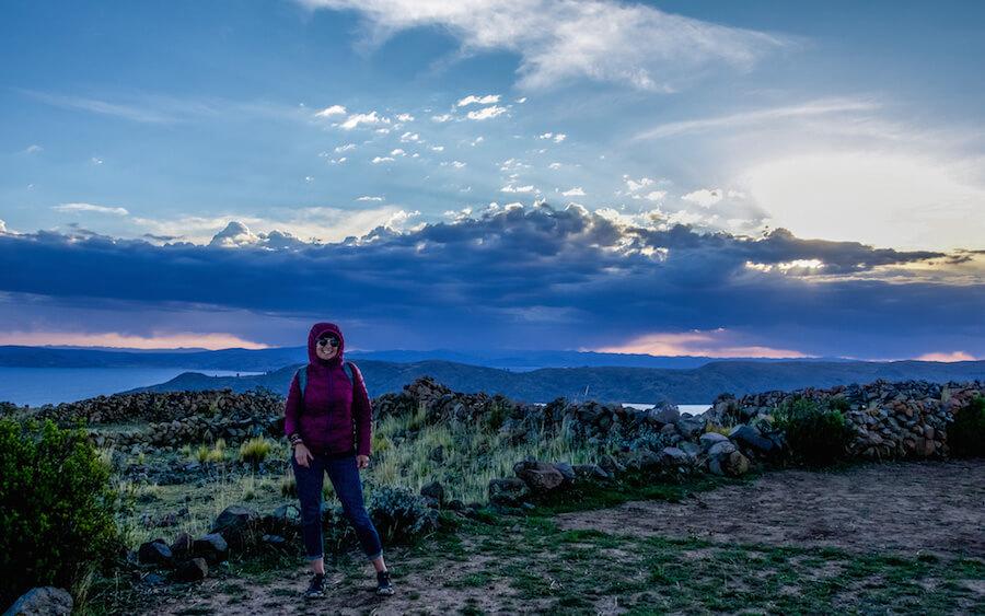 Solo Travel in South America: lady in burgundy jacket with hood up,standing wiht a view over a lake and mountains and a pink sunset