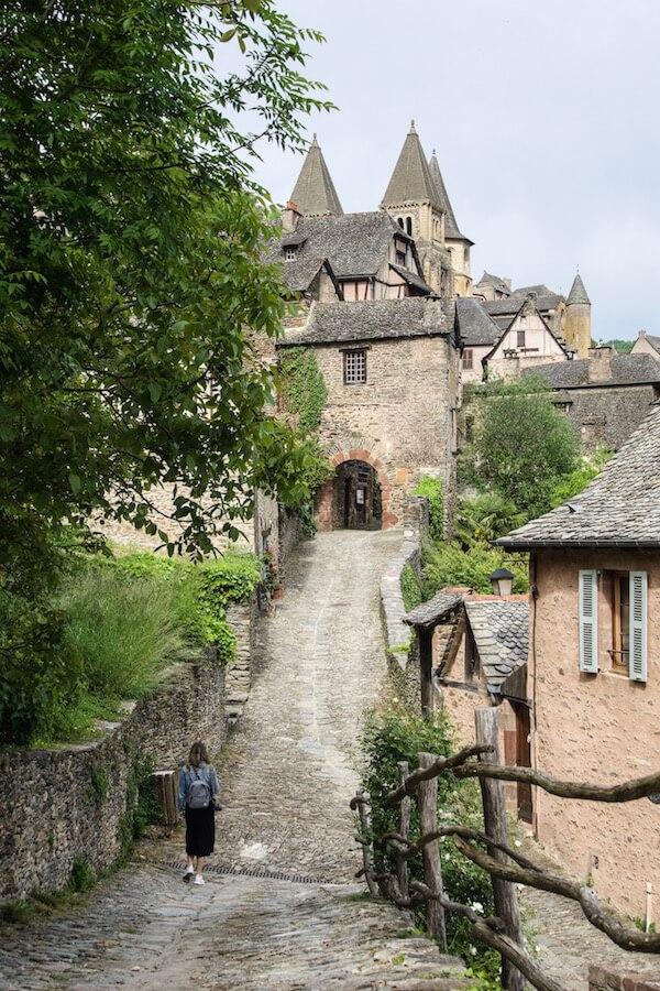 Conques = girl walking towards and stone arched gateway into the village