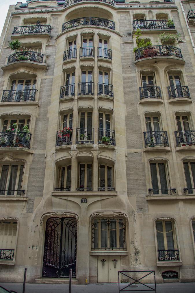 Art Nouveau in Paris 16: Looking at 4 stories of beige toned building,  Window balconies are stunning wrought iron designs. Shrubs and flowers on some of the baclonies