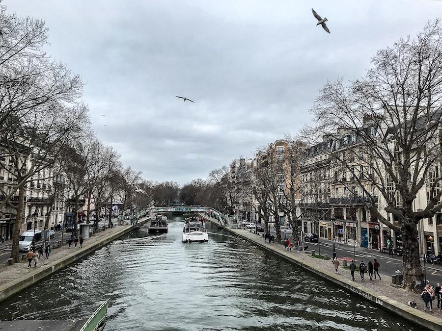 Canal Saint-Martin Paris: 2 boats passing, birds overhead, bare trees, people walking on either side of the canal