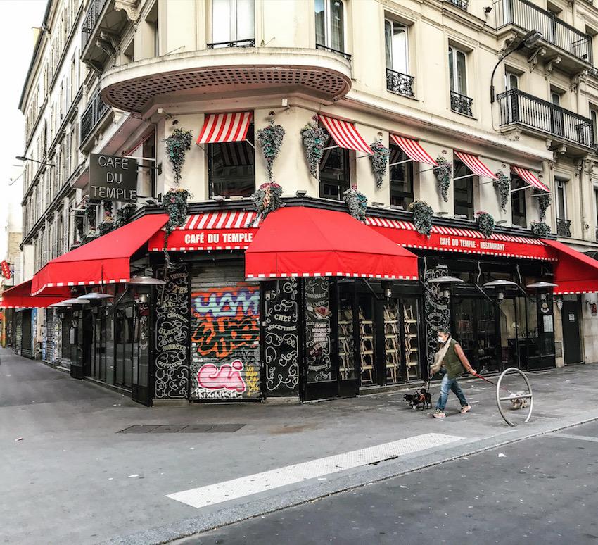 Paris during Coronavirus: A closed café with red and white striped awnings. A masked lady walks her two dogs